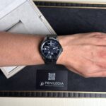 Wrist with a dark blue dial luxury watch (Ulysse Nardin) on a metal bracelet, beside a beige display box and a Privlegia card.