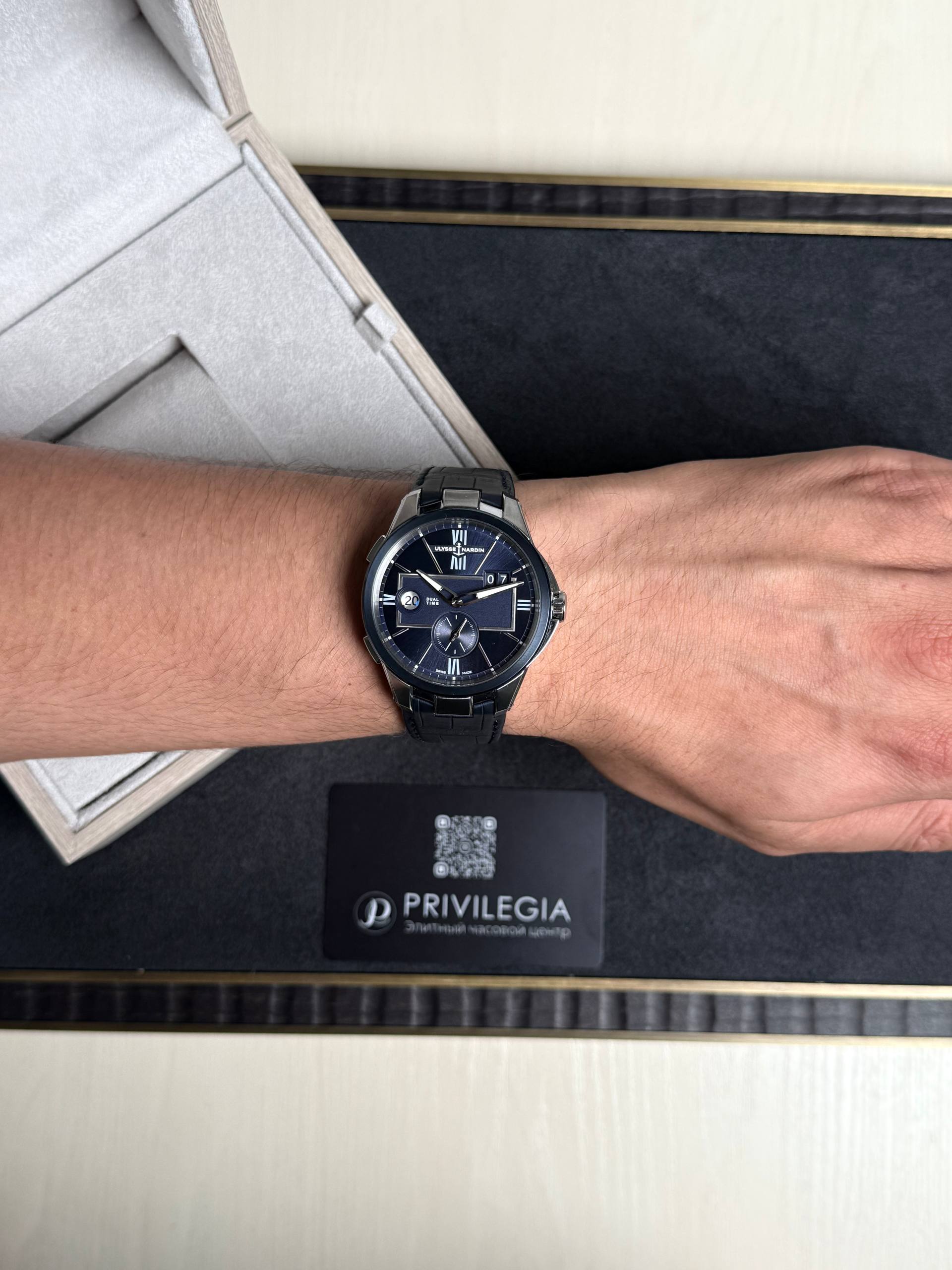 Wrist with a dark blue dial luxury watch (Ulysse Nardin) on a metal bracelet, beside a beige display box and a Privlegia card.