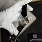 Back of a mechanical wristwatch with a visible rotor, held by a gloved hand in white gloves