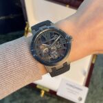 Skeleton-dial wristwatch with visible gears, dark case and gray strap on a knit-sleeved wrist; display box in background.