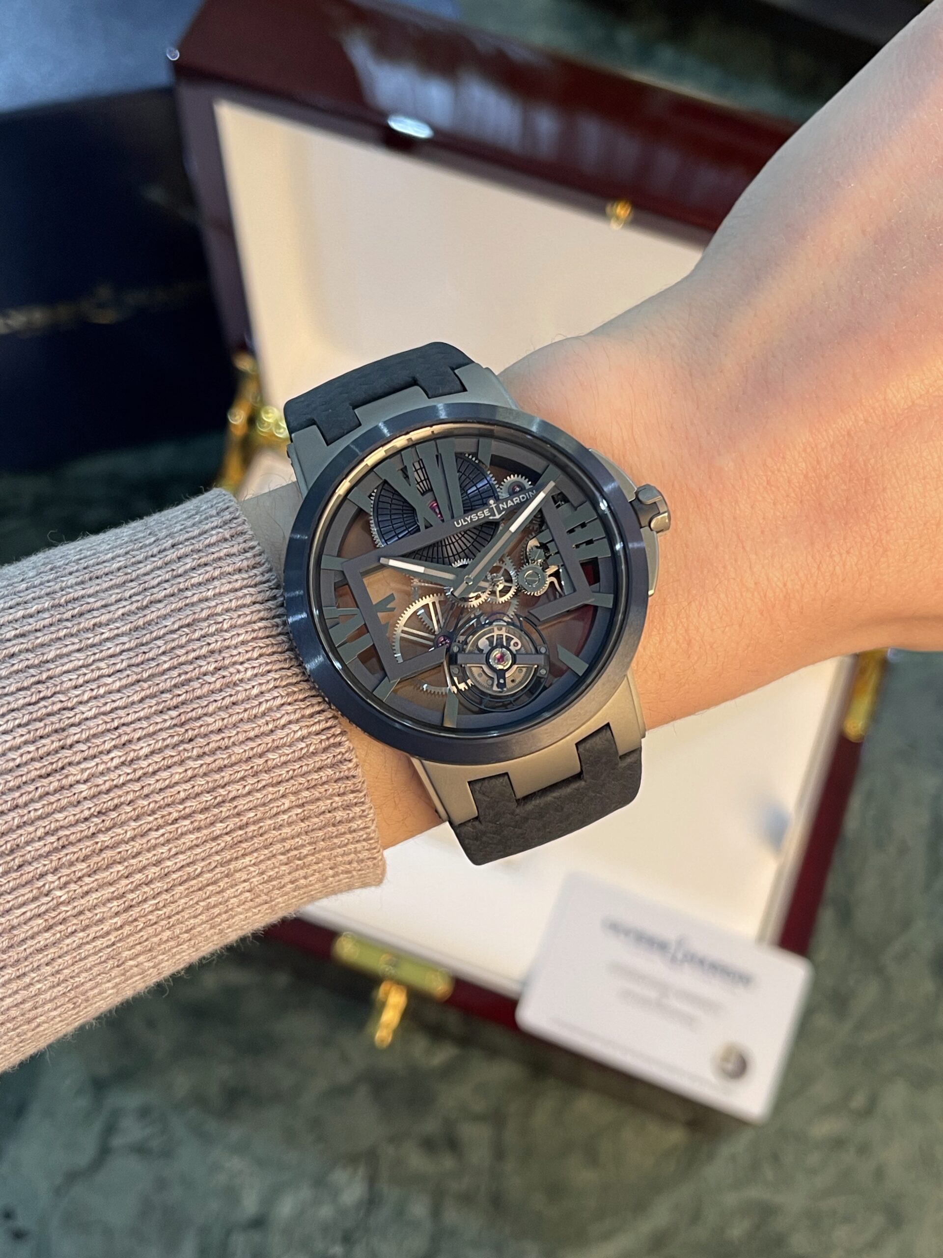 Skeleton-dial wristwatch with visible gears, dark case and gray strap on a knit-sleeved wrist; display box in background.