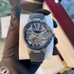 Skeleton dial wristwatch with blue-gray case and strap held by gloved hand, display box in background