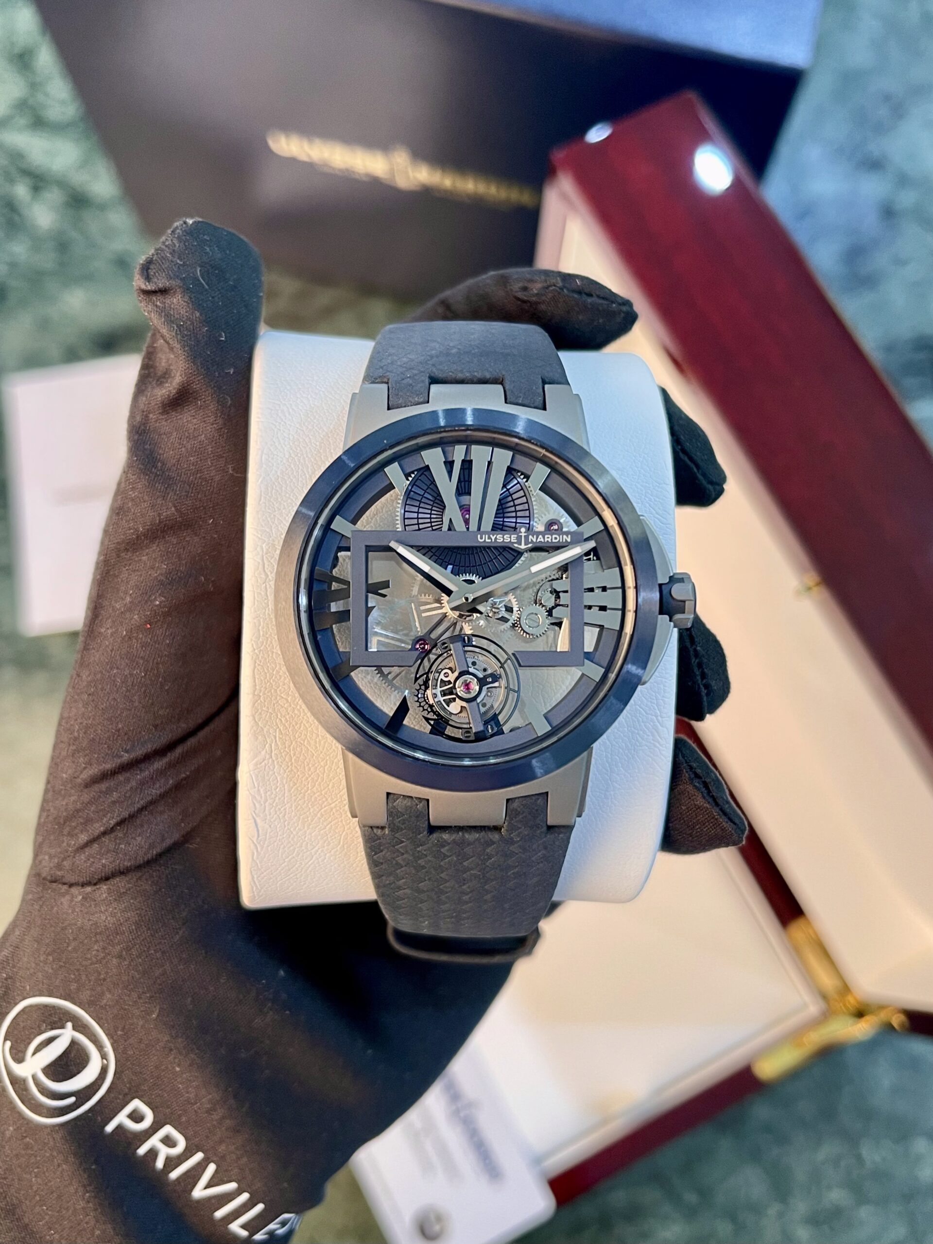 Skeleton dial wristwatch with blue-gray case and strap held by gloved hand, display box in background