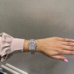 Wrist wearing a diamond-studded silver watch with a pale pink satin sleeve showing on the left arm.