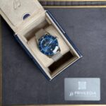 Blue dial Breitling watch in a beige suede presentation box with metal mesh strap, displayed on a dark surface near a Privilegia card.