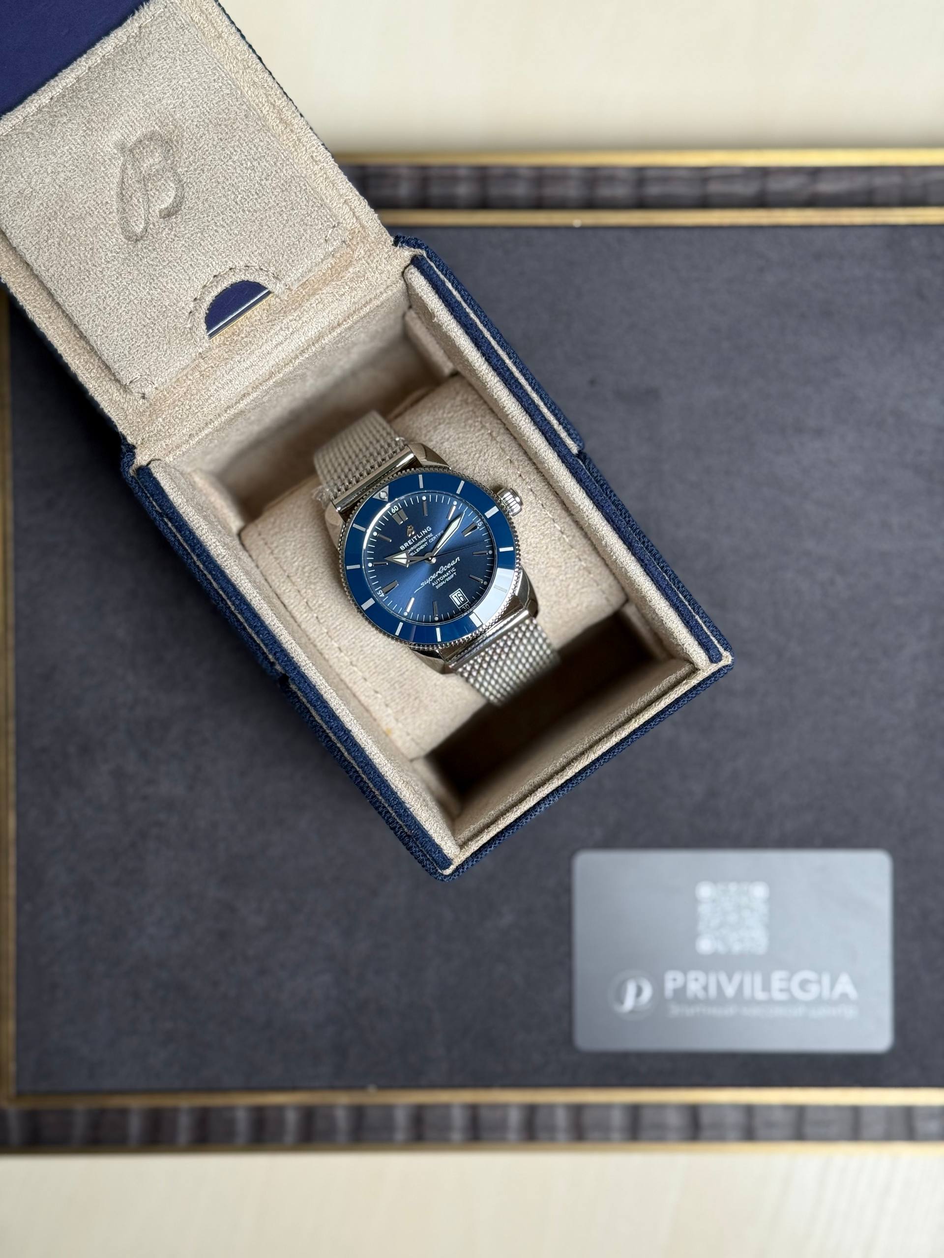 Blue dial Breitling watch in a beige suede presentation box with metal mesh strap, displayed on a dark surface near a Privilegia card.