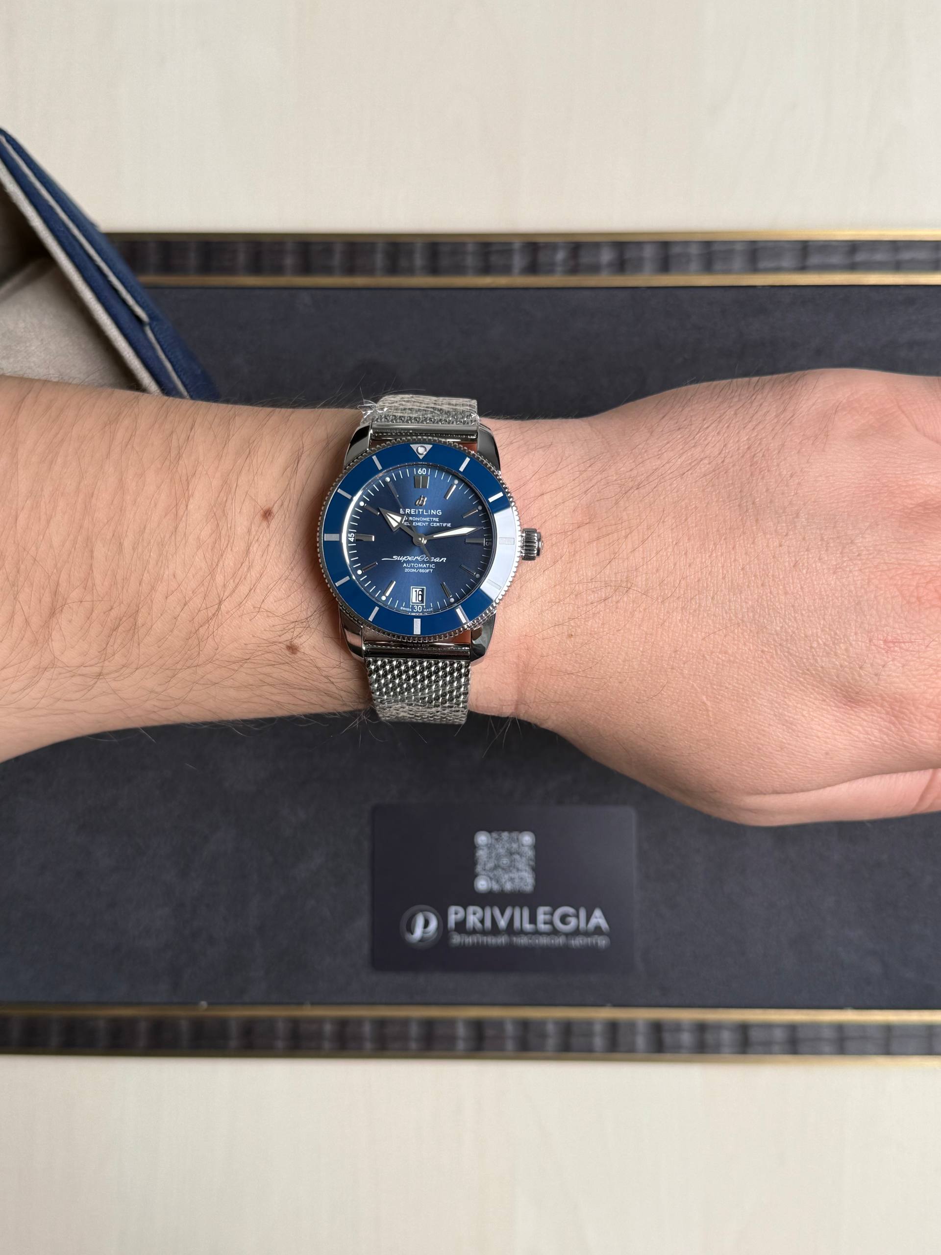 Breitling Superocean blue-dial wristwatch with stainless mesh band on the wrist, shown close-up on a dark background with a Privilegia card nearby.