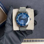 Breitling SuperOcean automatic watch with blue dial and mesh metal bracelet on a beige display pillow held by a gloved hand.