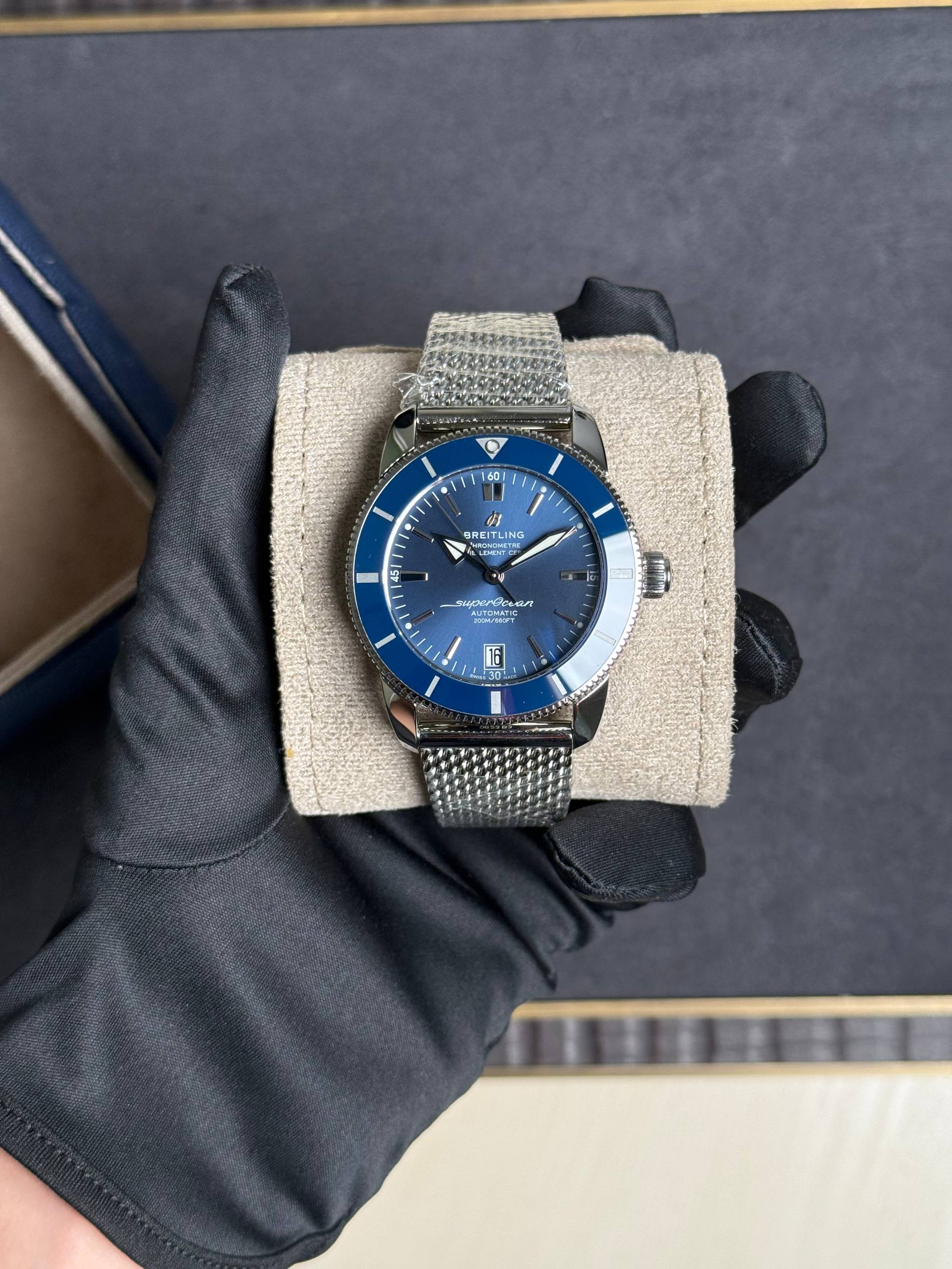Breitling SuperOcean automatic watch with blue dial and mesh metal bracelet on a beige display pillow held by a gloved hand.