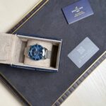 Breitling wristwatch with blue dial in an open beige box on a dark surface, mesh bracelet visible next to a gray card with a QR code.