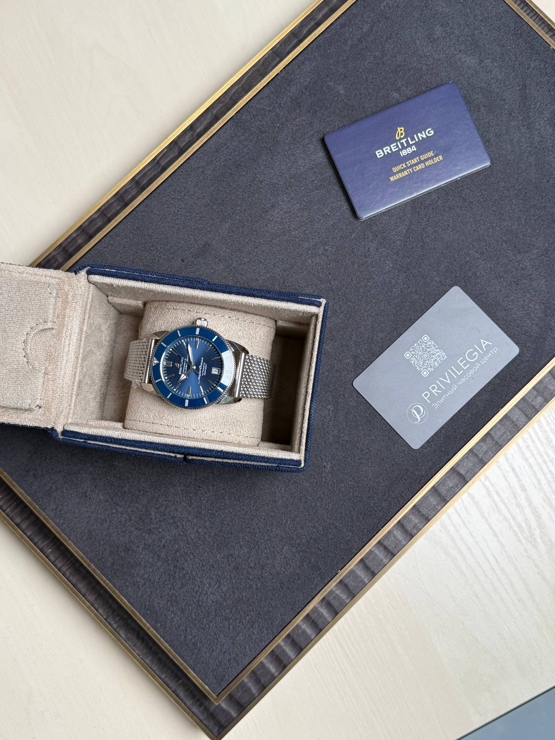 Breitling wristwatch with blue dial in an open beige box on a dark surface, mesh bracelet visible next to a gray card with a QR code.