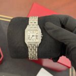 Diamond-encrusted silver Cartier wristwatch with Roman numerals on a white dial, resting on a black glove.