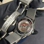 Close-up of a Breitling stainless-steel chronograph watch with a mesh bracelet, resting on dark fabric; the transparent case back reveals the inner mechanism.