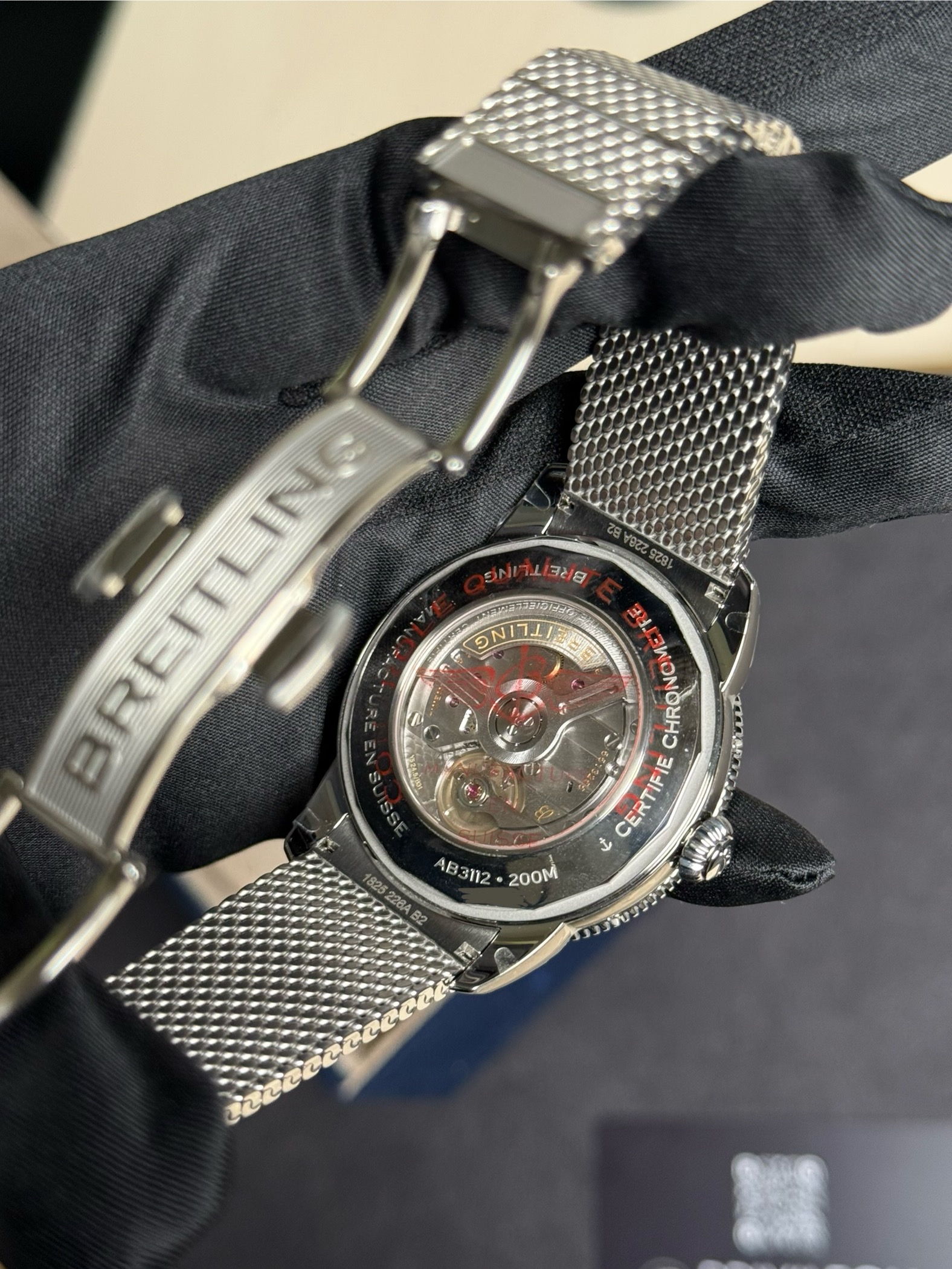 Close-up of a Breitling stainless-steel chronograph watch with a mesh bracelet, resting on dark fabric; the transparent case back reveals the inner mechanism.