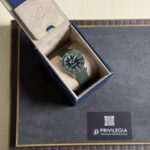 Green metal-mesh wristwatch in an open blue presentation box with beige interior. A Privilegia card sits on a dark display mat nearby.