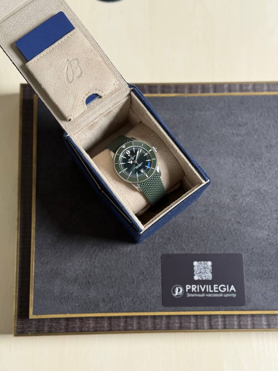 Green metal-mesh wristwatch in an open blue presentation box with beige interior. A Privilegia card sits on a dark display mat nearby.