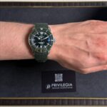 Breitling wristwatch with a dark dial and green fabric strap on a forearm resting on a black display box labeled Privilegia.
