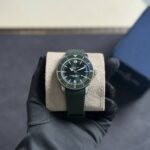 Green Breitling wristwatch with a textured green strap resting on a beige cushion, held by a gloved hand.