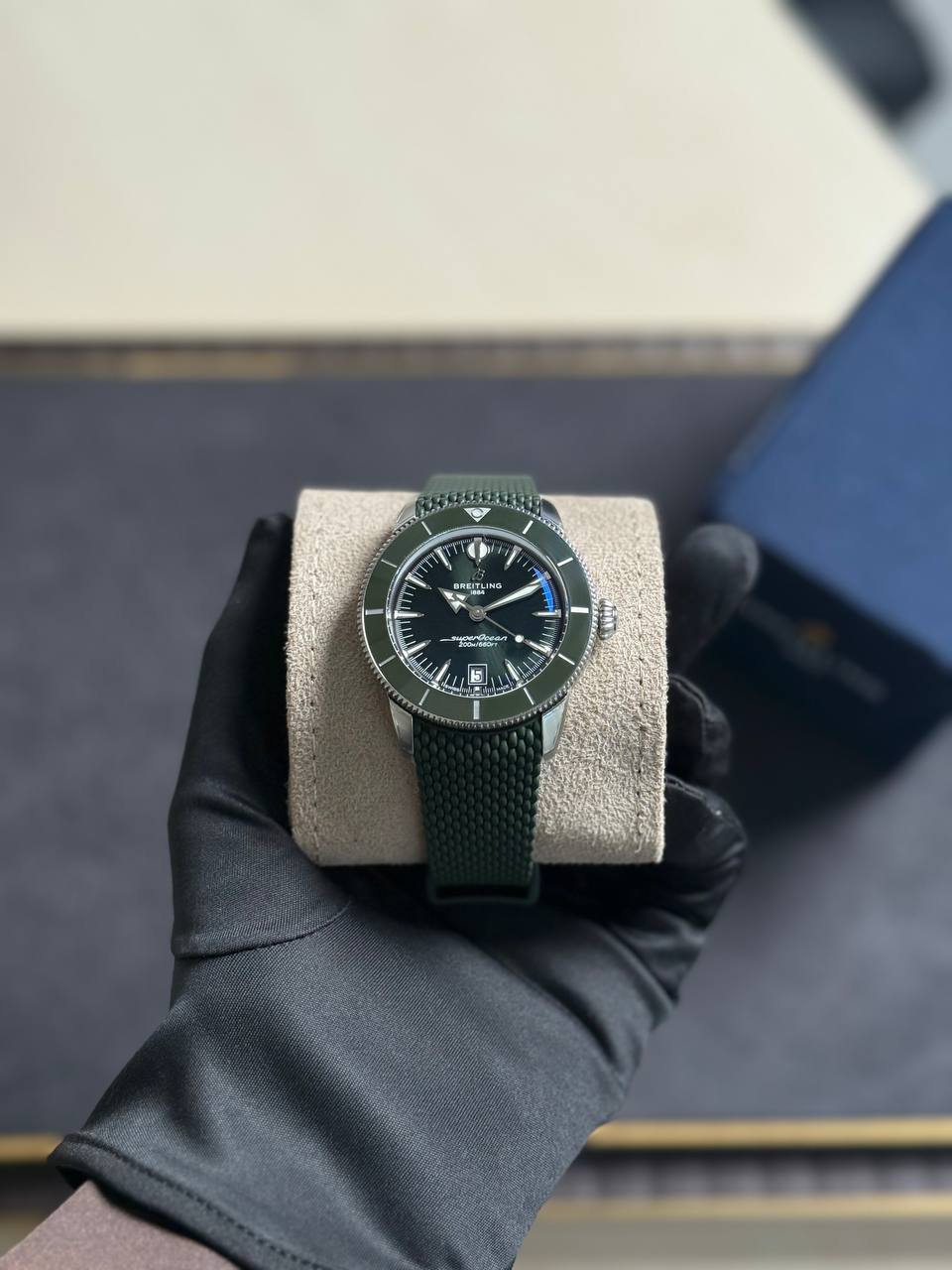 Green Breitling wristwatch with a textured green strap resting on a beige cushion, held by a gloved hand.