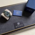 Breitling watch with green strap on a beige cushion, next to a blue warranty card and a navy Breitling box on a black tray.
