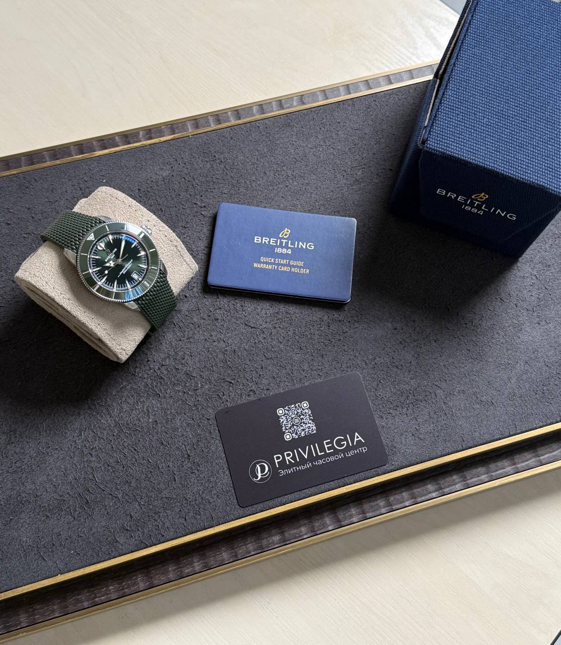 Breitling watch with green strap on a beige cushion, next to a blue warranty card and a navy Breitling box on a black tray.