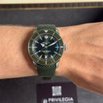 Breitling SuperOcean wristwatch with a green bezel and green mesh strap on a forearm, with a Privilegia box in the blurred background.