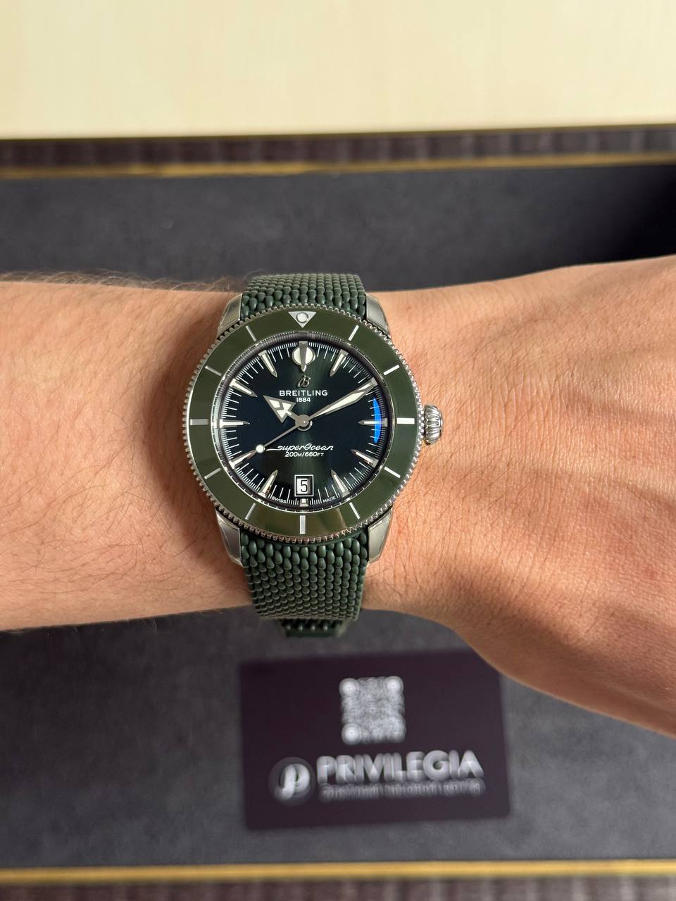 Breitling SuperOcean wristwatch with a green bezel and green mesh strap on a forearm, with a Privilegia box in the blurred background.