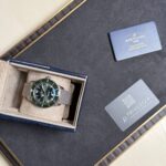 Breitling wristwatch with a metal mesh band in a beige padded box on a dark surface, with a blue warranty card nearby.