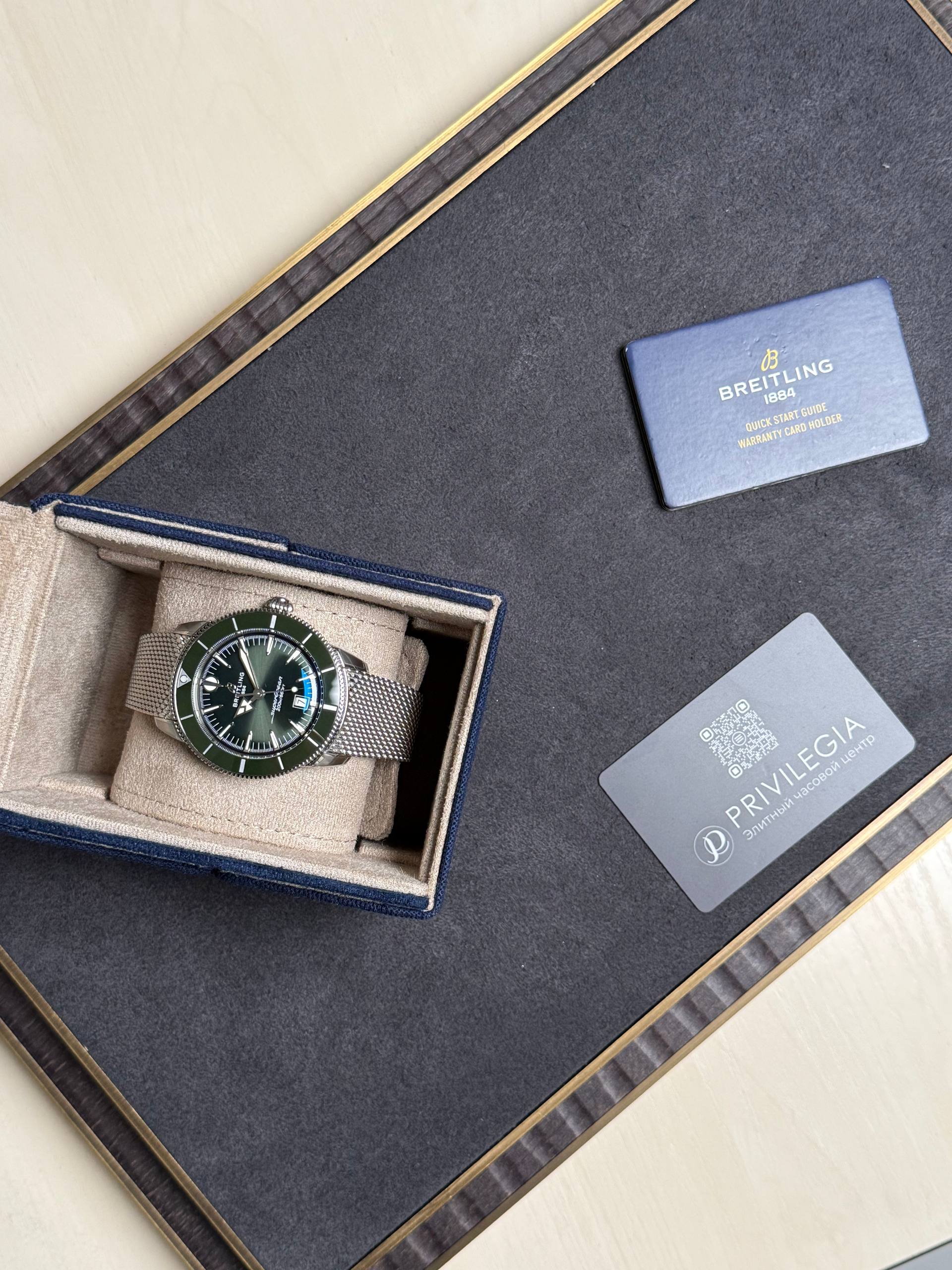 Breitling wristwatch with a metal mesh band in a beige padded box on a dark surface, with a blue warranty card nearby.