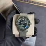 Breitling SuperOcean wristwatch with a green bezel and mesh stainless steel bracelet, held by gloved hands over a beige display cushion.