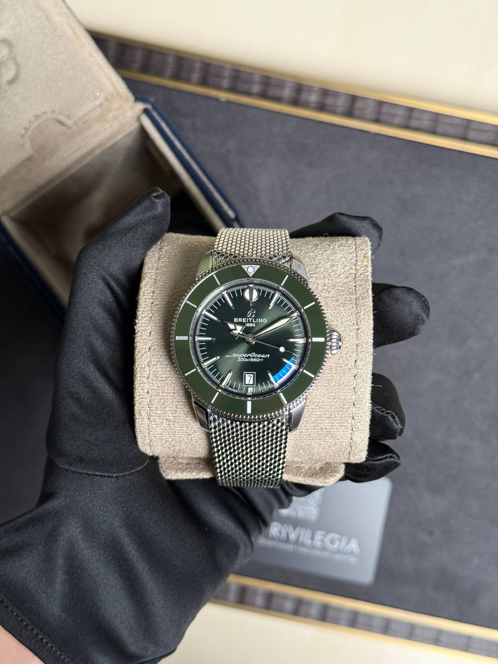 Breitling SuperOcean wristwatch with a green bezel and mesh stainless steel bracelet, held by gloved hands over a beige display cushion.