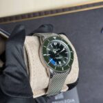 Close-up of a Breitling SuperOcean diver watch with a green bezel on a gloved hand, metallic mesh band visible against dark background.