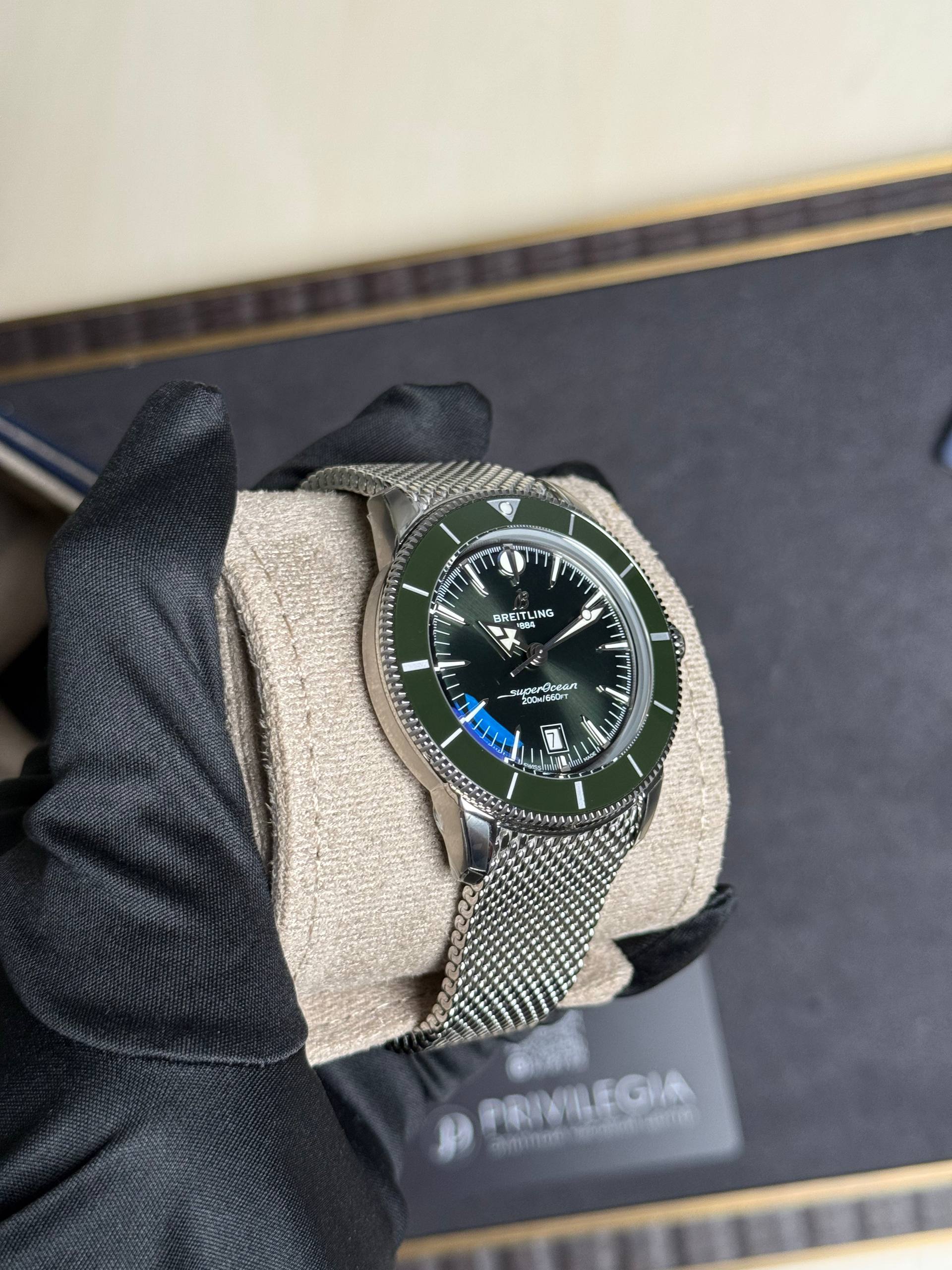 Close-up of a Breitling SuperOcean diver watch with a green bezel on a gloved hand, metallic mesh band visible against dark background.