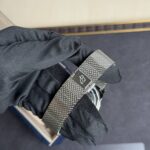 Close-up of a wrist wearing a silver mesh metal watch band over a dark fabric sleeve. The watch face is partially visible at the bottom edge.