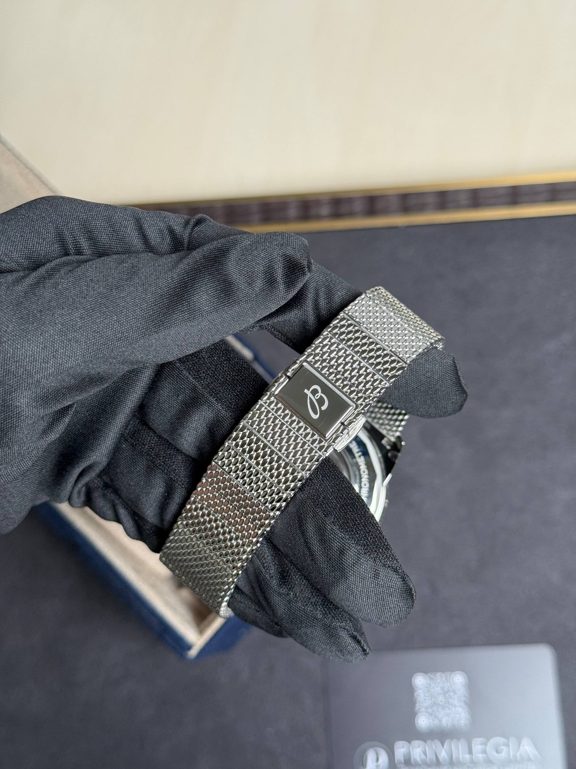 Close-up of a wrist wearing a silver mesh metal watch band over a dark fabric sleeve. The watch face is partially visible at the bottom edge.