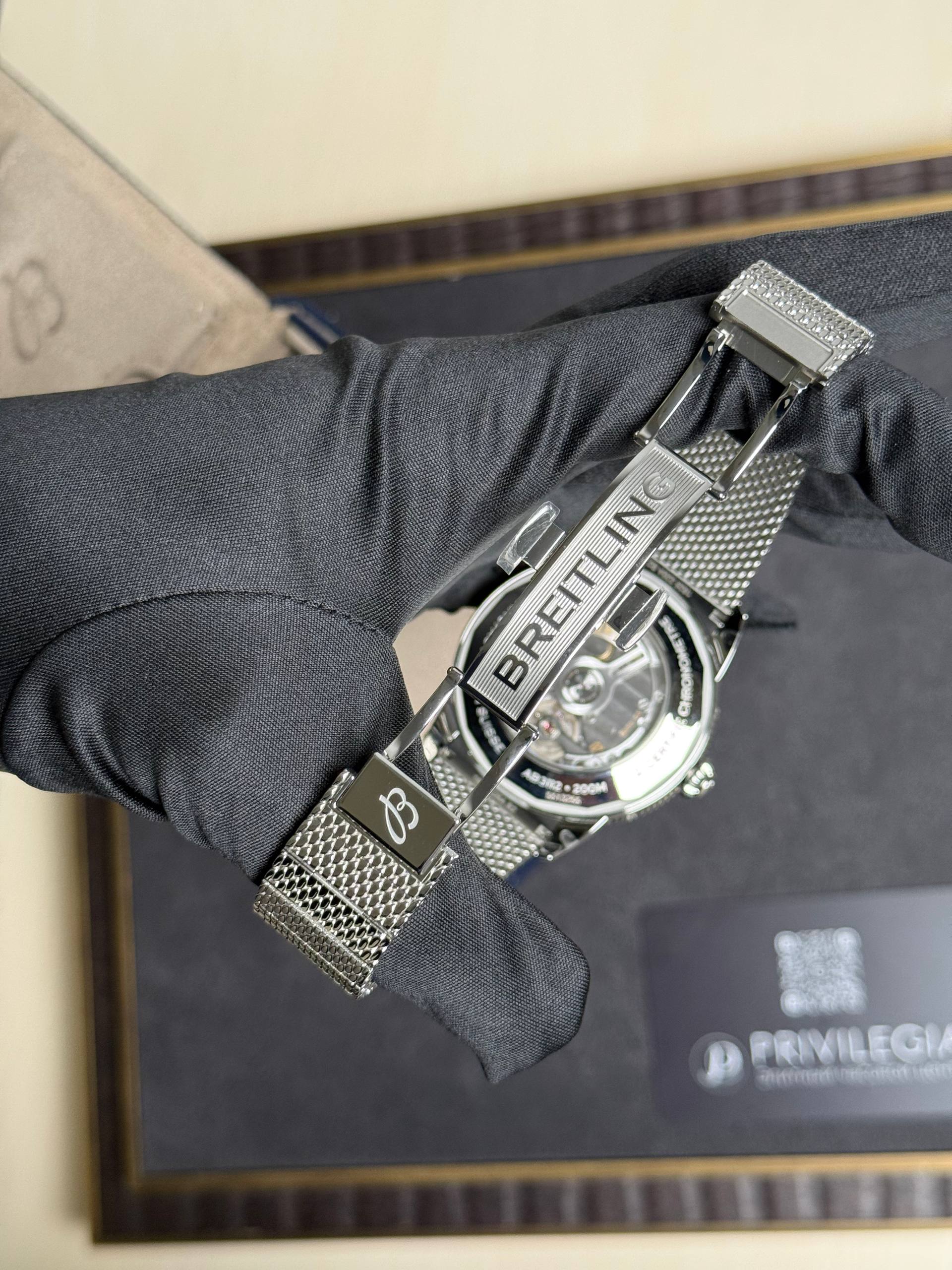 Close-up of a Breitling watch with a metal mesh bracelet on a gloved wrist, showing the clasp and logo text.