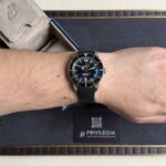 Close-up of a Breitling diver's watch on a forearm, featuring a black dial, silver bezel, and black textured strap.
