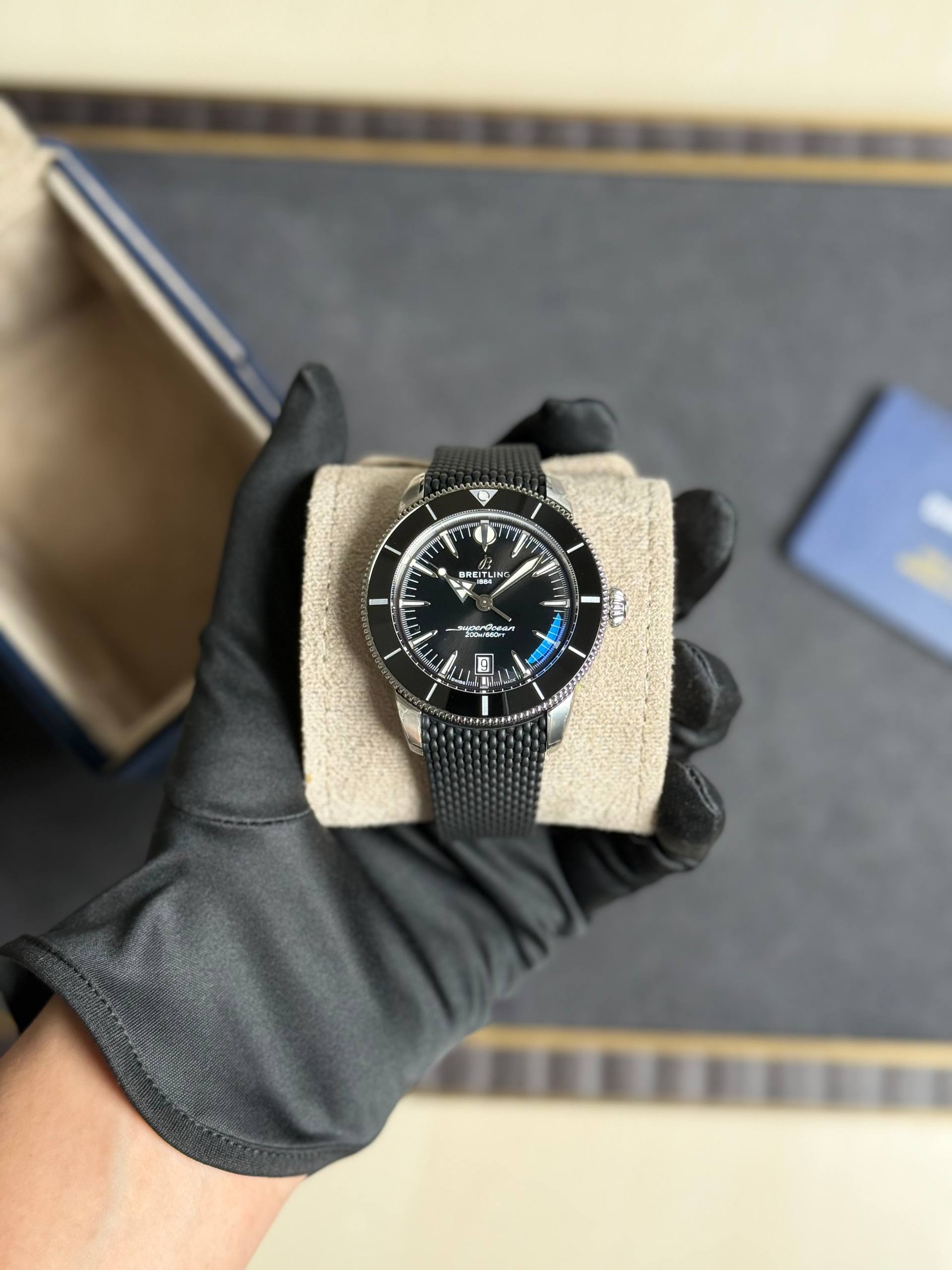 Breitling wristwatch with a black dial and mesh strap held on a beige display cushion by a gloved hand.