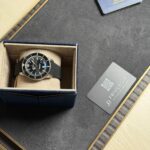 Breitling wristwatch in blue velvet box with beige interior, placed on a textured surface beside a warranty card and privacy card.