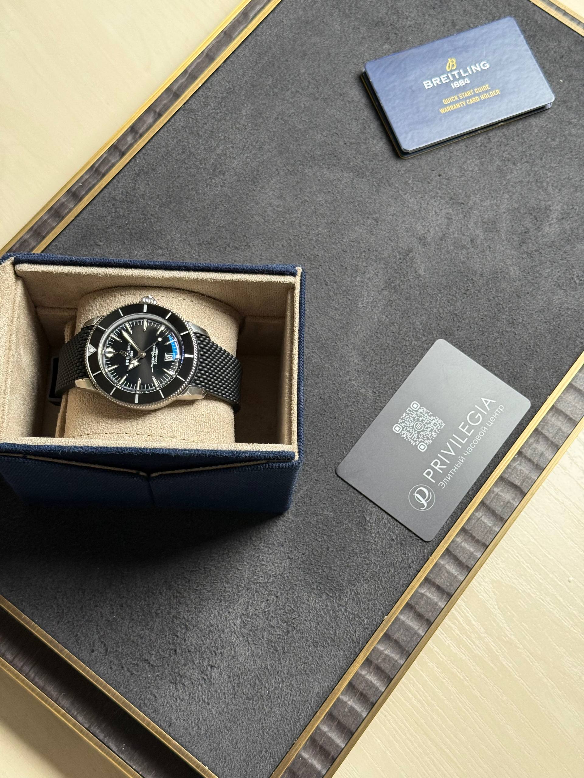 Breitling wristwatch in blue velvet box with beige interior, placed on a textured surface beside a warranty card and privacy card.