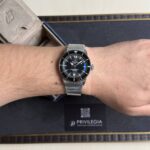 Wrist with a silver Breitling Superocean dive watch and mesh bracelet, on a dark display box background.