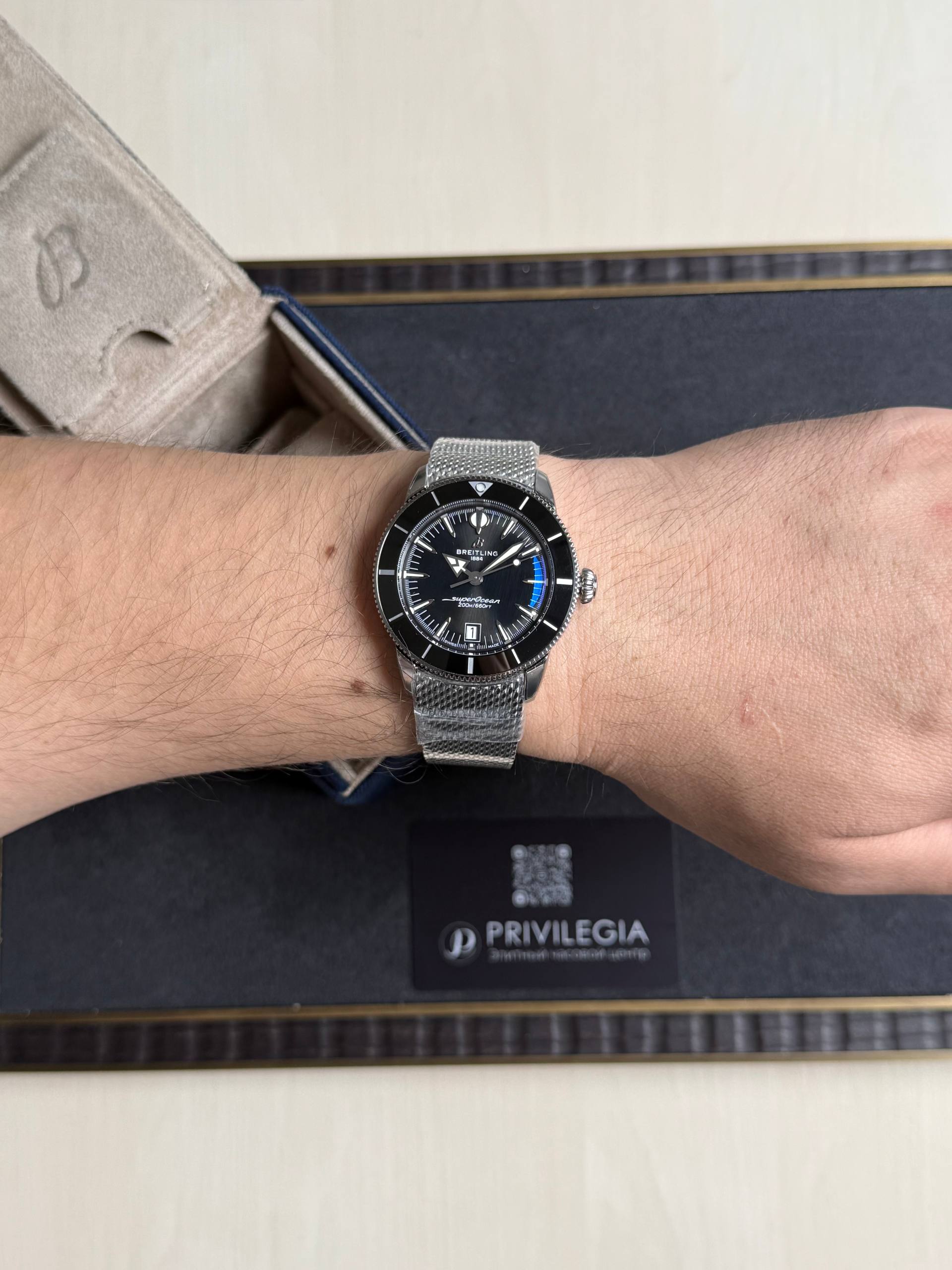 Wrist with a silver Breitling Superocean dive watch and mesh bracelet, on a dark display box background.