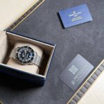 Breitling wristwatch with a metal mesh band in an open beige-lined box on a dark display surface, next to a blue warranty card labeled Breitling 1884 and a Privilegia card.