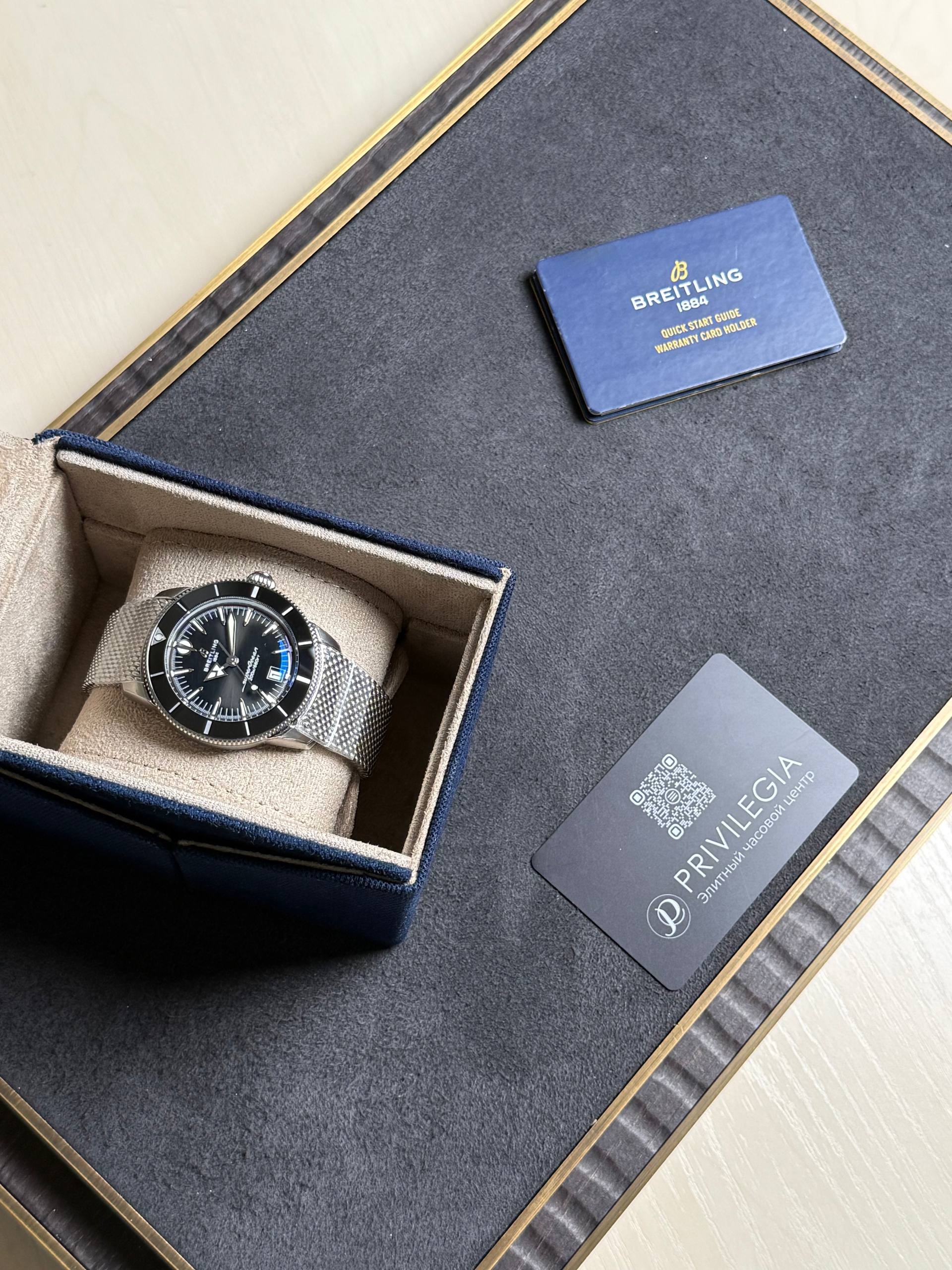 Breitling wristwatch with a metal mesh band in an open beige-lined box on a dark display surface, next to a blue warranty card labeled Breitling 1884 and a Privilegia card.