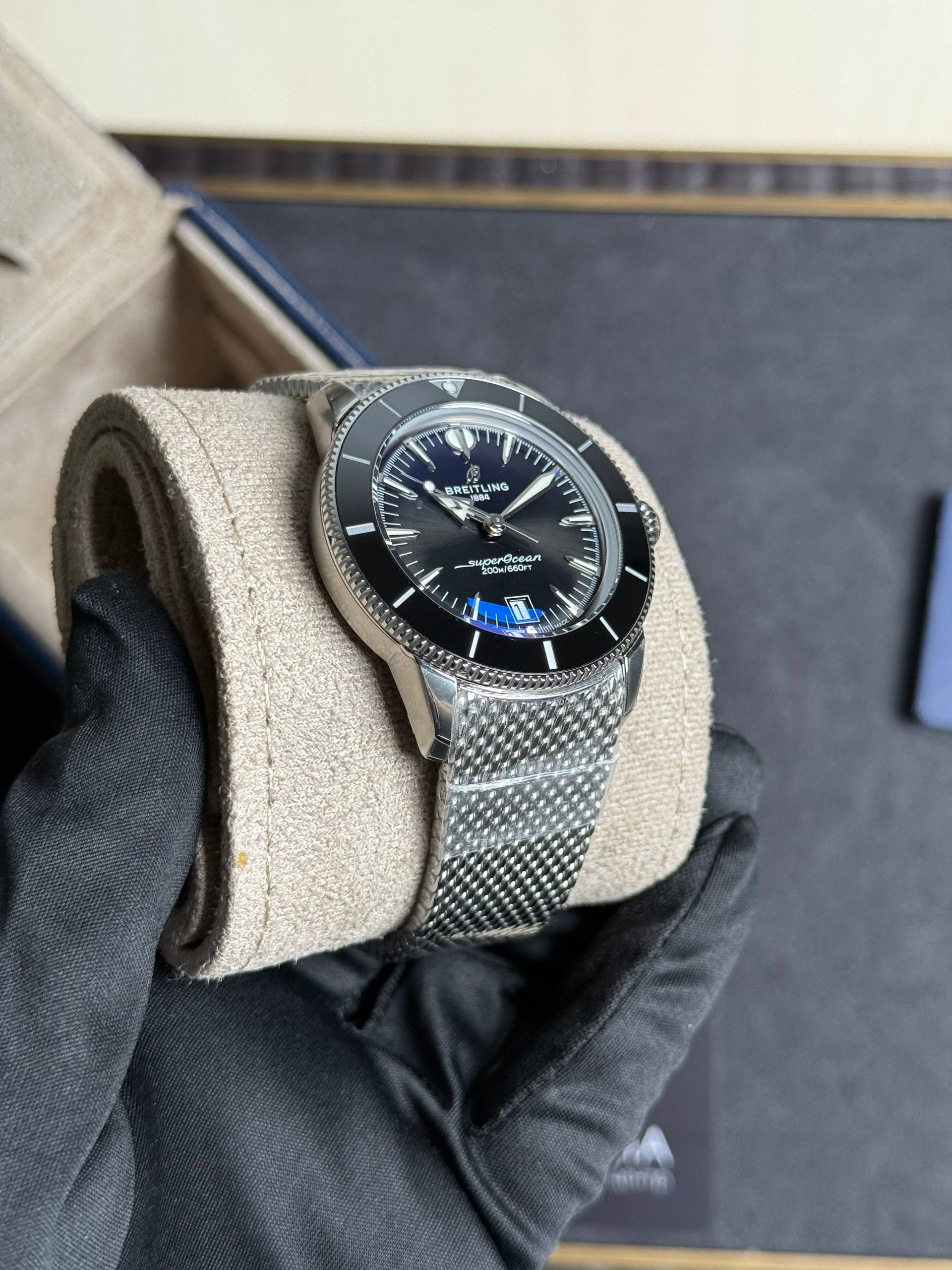 Breitling SuperOcean wristwatch with black dial, silver bezel, and mesh bracelet resting on a beige cushion in a gloved hand.