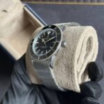 Close-up of a stainless steel Breitling SuperOcean watch on a metal mesh bracelet, held by gloved hands against a neutral background.