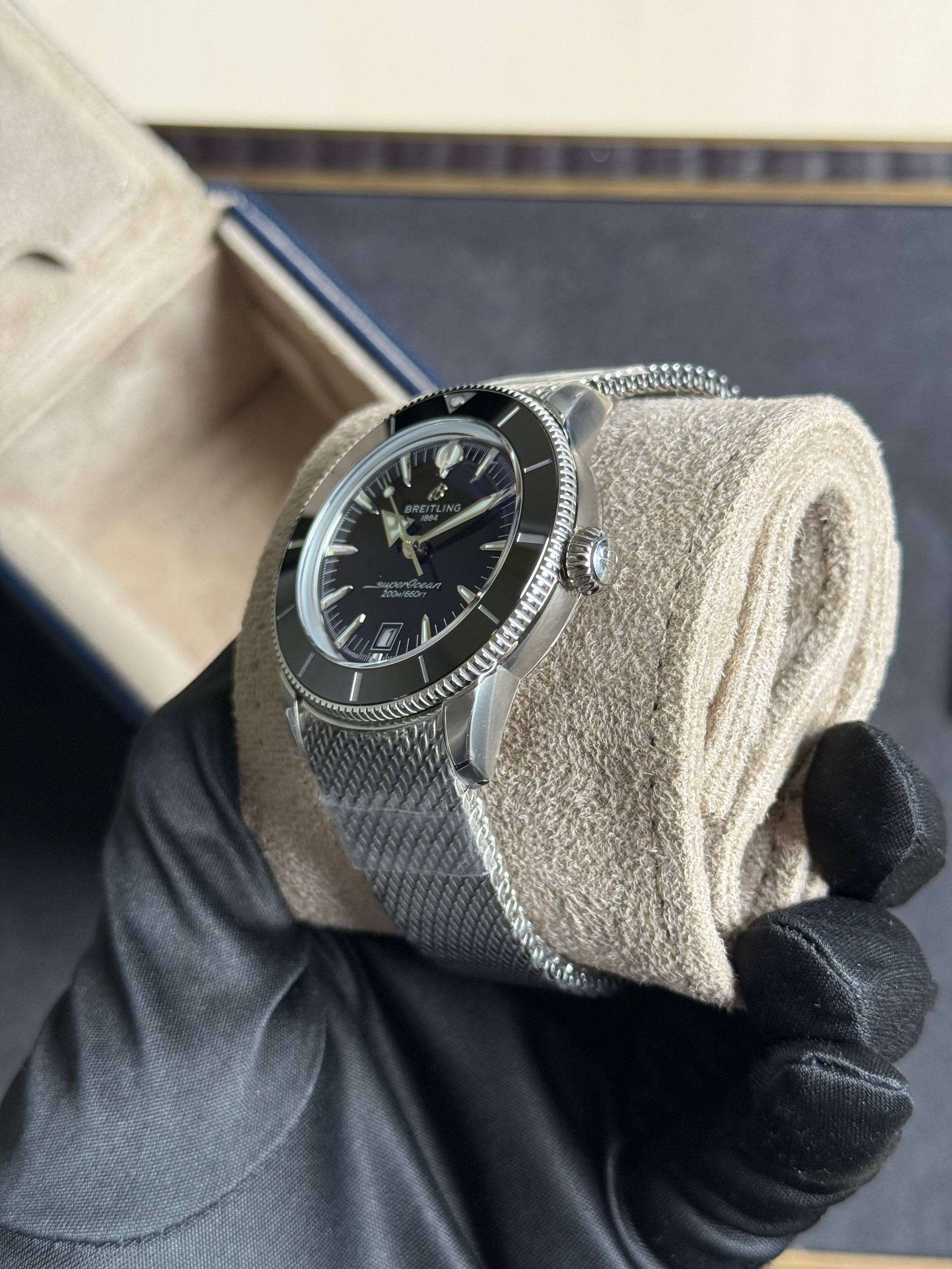 Close-up of a stainless steel Breitling SuperOcean watch on a metal mesh bracelet, held by gloved hands against a neutral background.