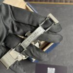 Close-up of a silver Breitling watch bracelet clasp on a black glove, with blurred box in the background.