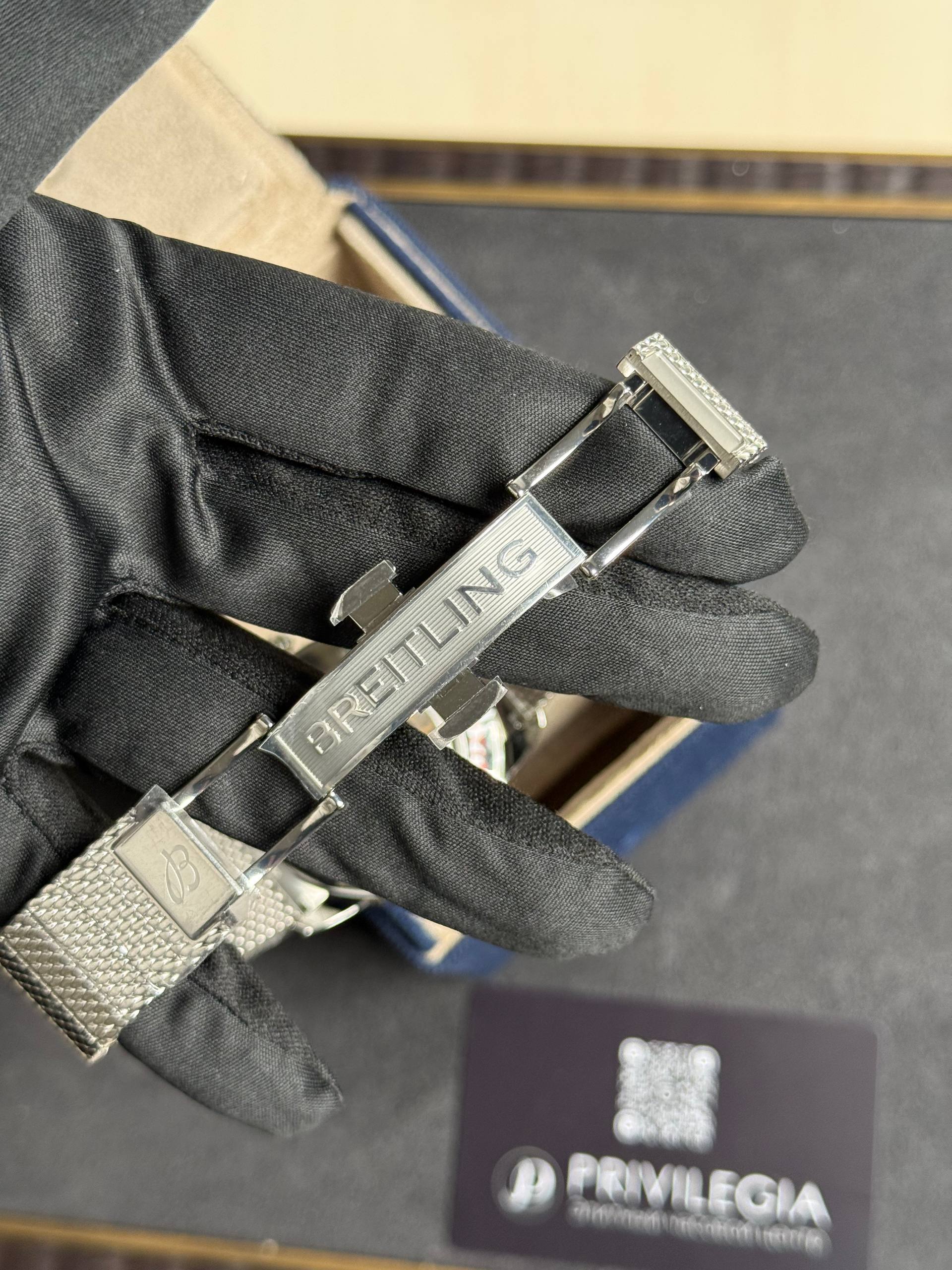 Close-up of a silver Breitling watch bracelet clasp on a black glove, with blurred box in the background.