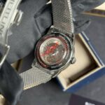 Back of a Breitling wristwatch with a metal mesh band resting on a gloved hand; you can see the red accent and engravings on the case back, including 'BREITLING' and 'MANUFACTURE'.